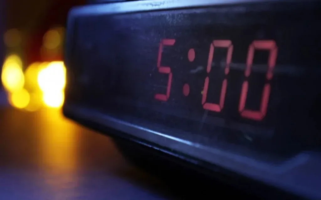 Digital alarm clock glowing red at 5:00 in the dark with warm blurred lights behind