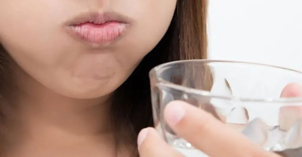 Close-up of a woman gargling with cheeks puffed out, holding a small glass of water
