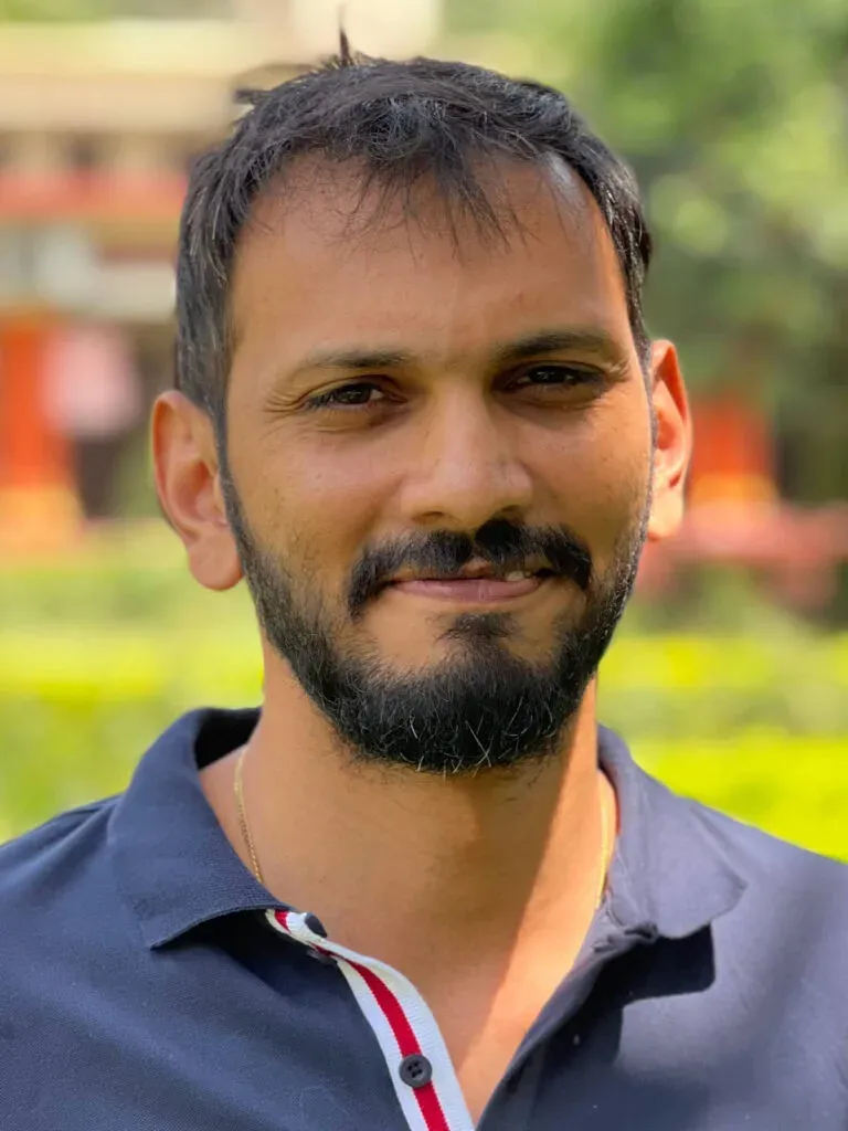 Smiling bearded man in a dark polo shirt outdoors with blurred greenery behind him