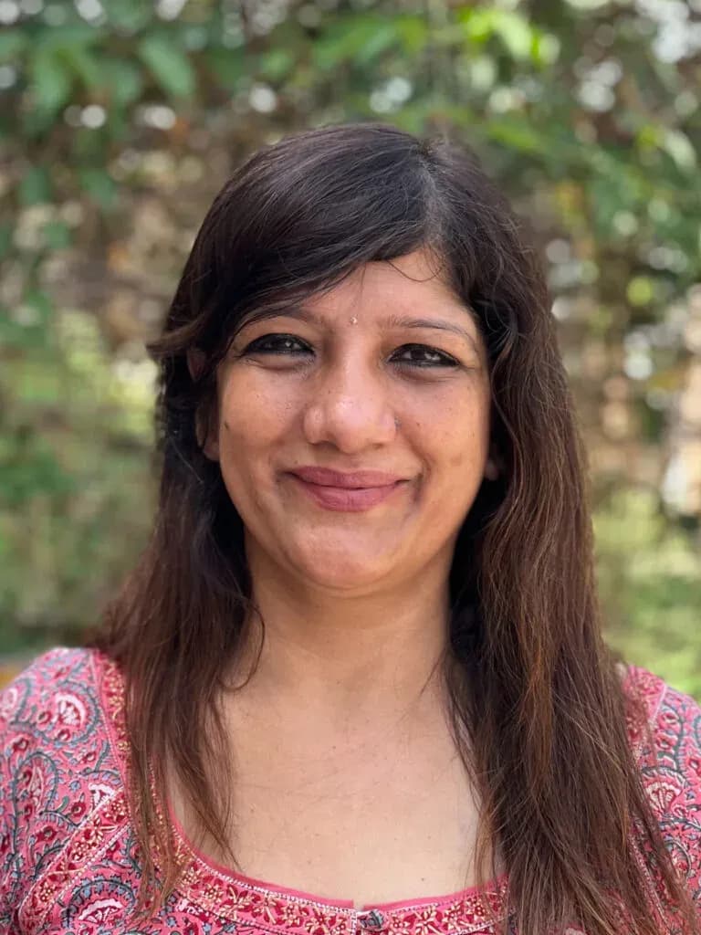 Smiling Indian woman with long dark hair in a pink printed top before leafy greenery