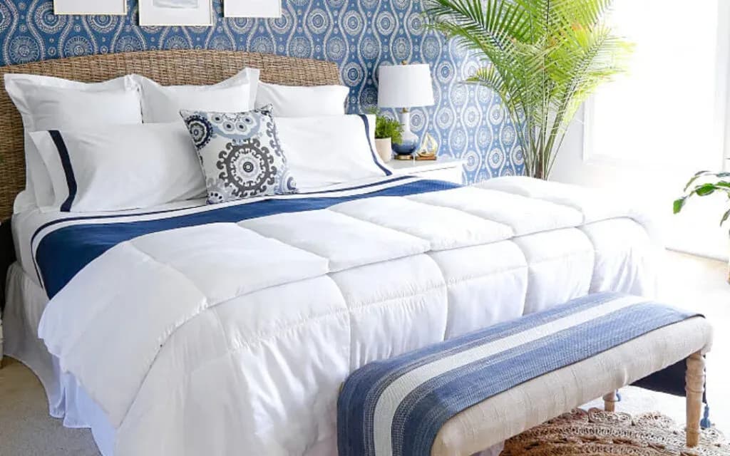 Neatly made bed with white bedding and navy trim beside blue patterned wallpaper and a palm
