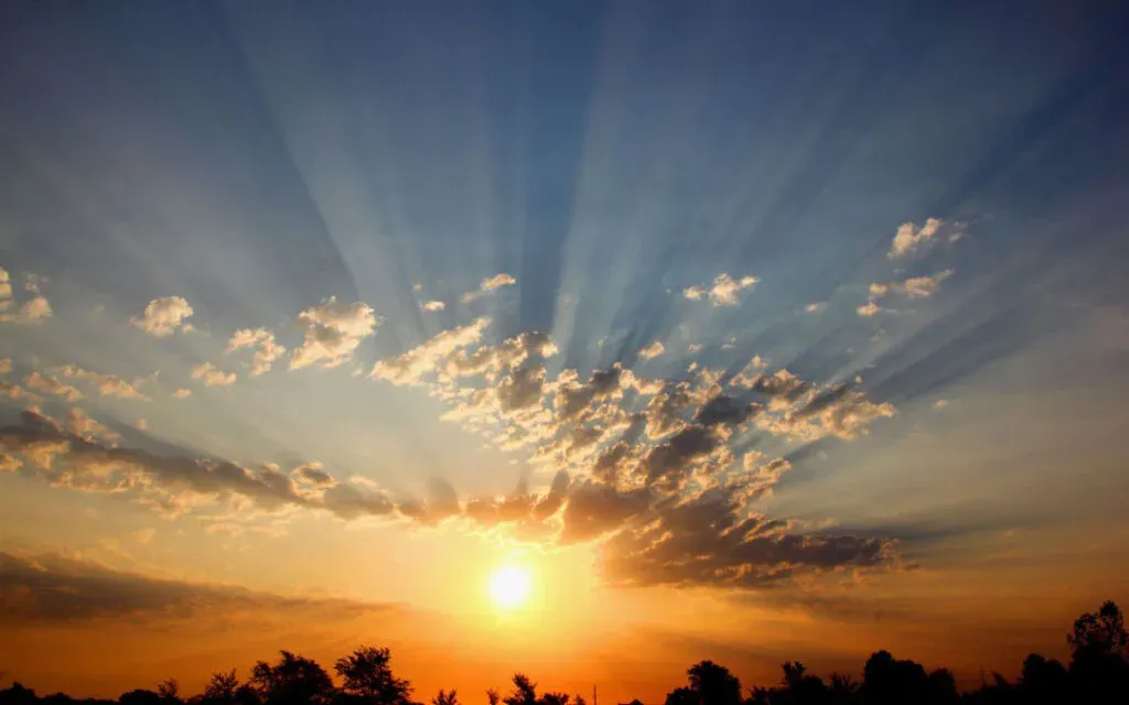Sun breaking through scattered clouds at sunset with rays fanning across the sky over silhouetted trees
