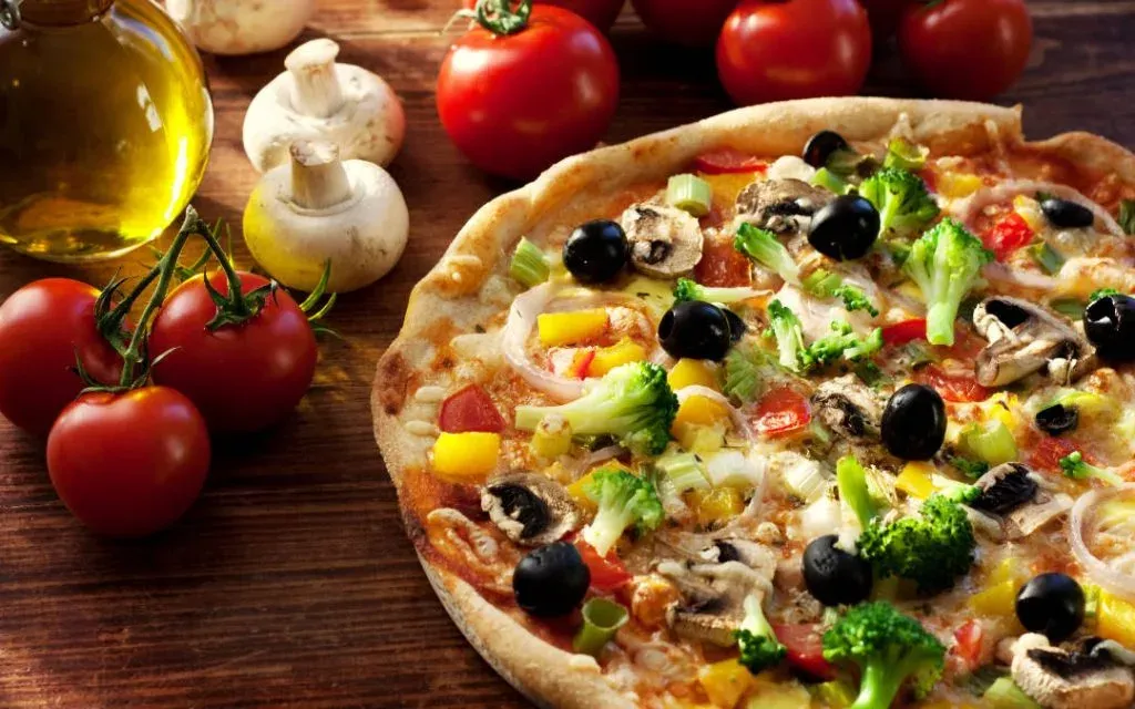 Vegetable pizza topped with olives, broccoli and mushrooms beside tomatoes and olive oil on wood