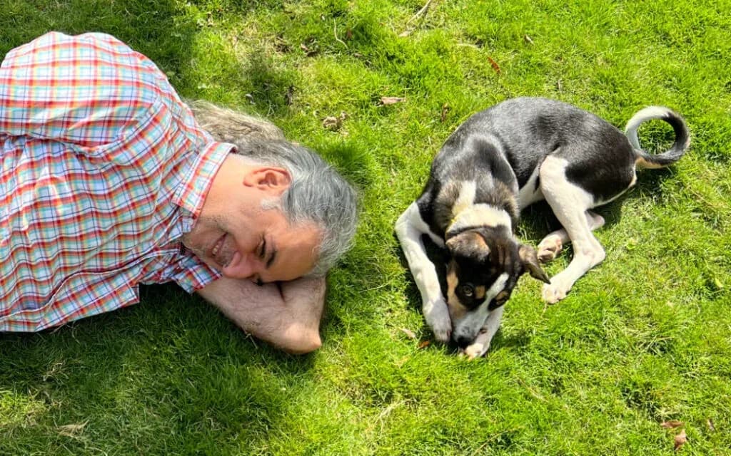 Grey-haired man lying on the grass smiling beside a black and white puppy