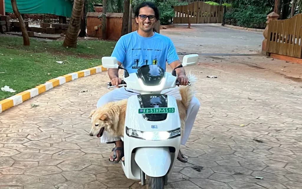 Dinesh smiling on a white electric scooter with a golden dog tucked at his feet