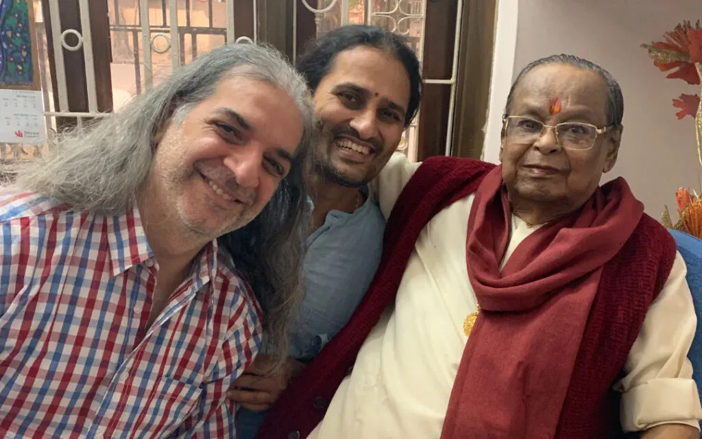 Two smiling men leaning close to a seated elderly man wearing a maroon shawl and tilak