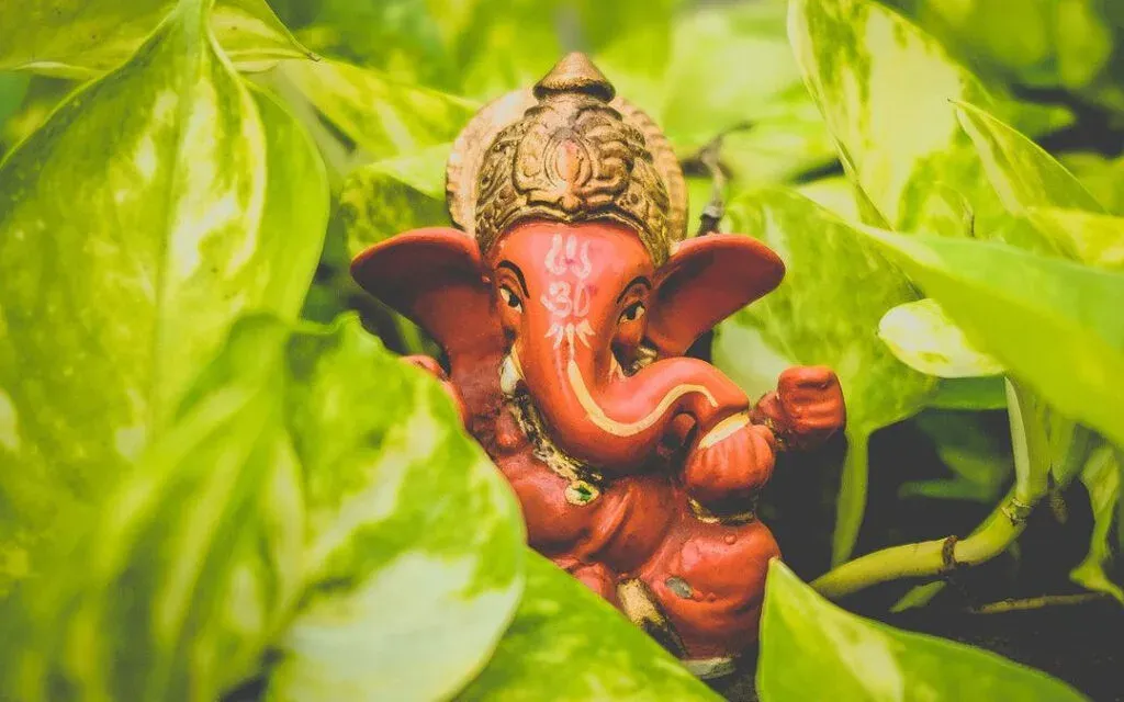A small red clay statue of Lord Ganesha peeking out among bright green pothos leaves