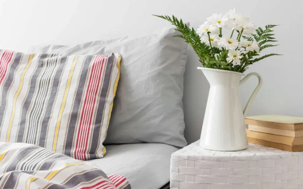 Grey striped cushions on a bed beside a white jug of daisies and stacked books