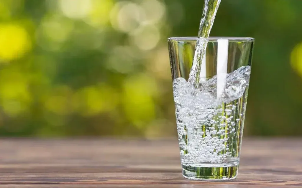 Water being poured into a clear glass on a wooden table against a leafy green background