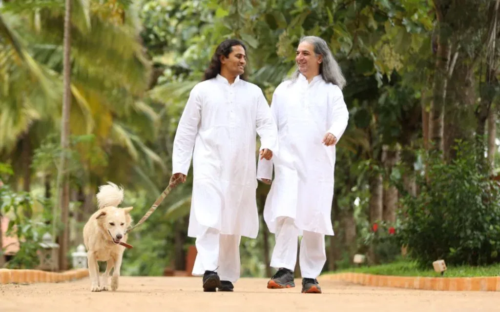 Two men in white kurtas walk a dog along a leafy garden path, smiling in conversation