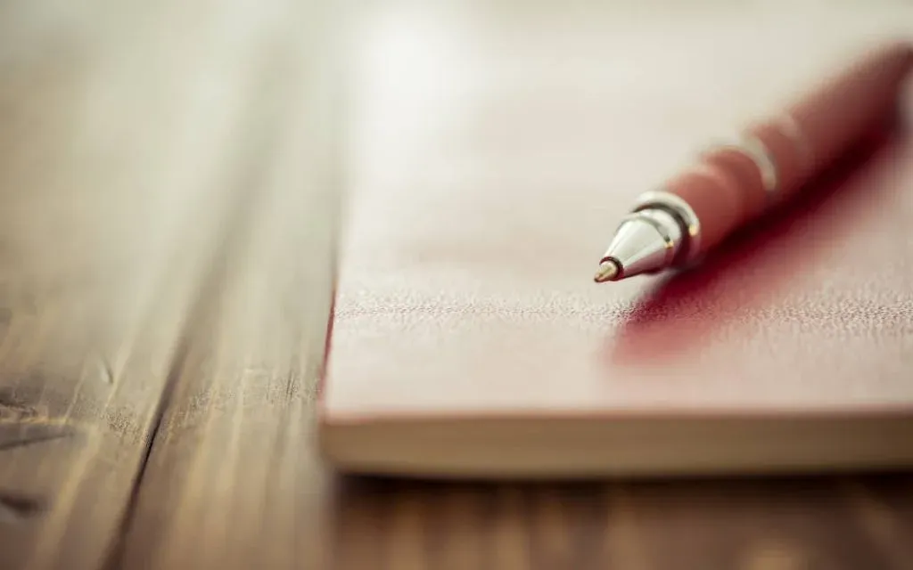 A red ballpoint pen resting on a closed red notebook on a wooden desk