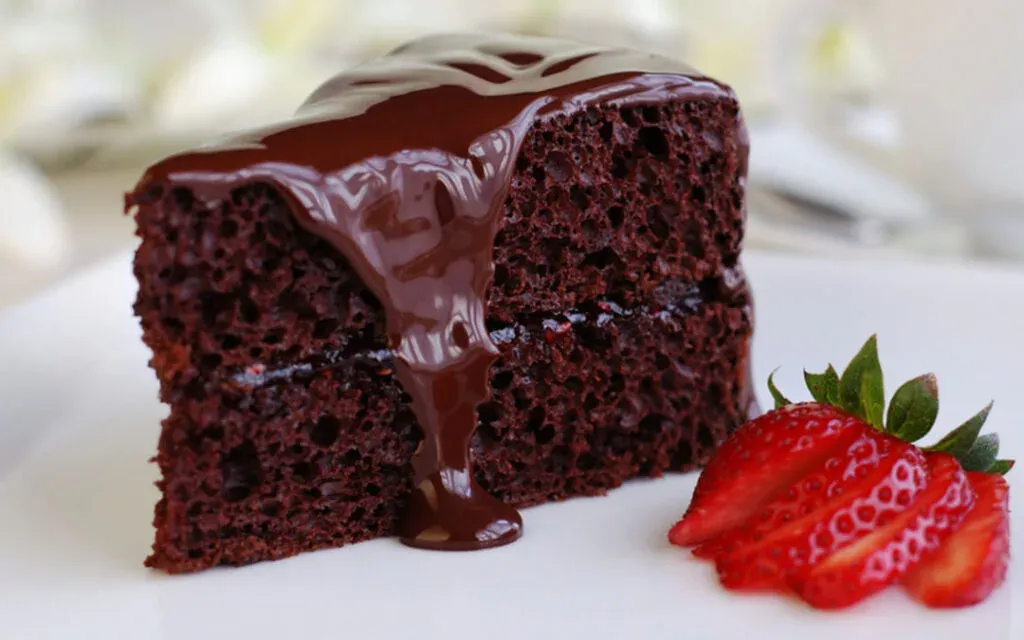Slice of chocolate cake with dripping ganache next to a fanned strawberry on a white plate