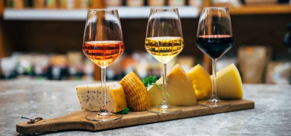 Three glasses of rose, white and red wine beside wedges of cheese on a wooden board