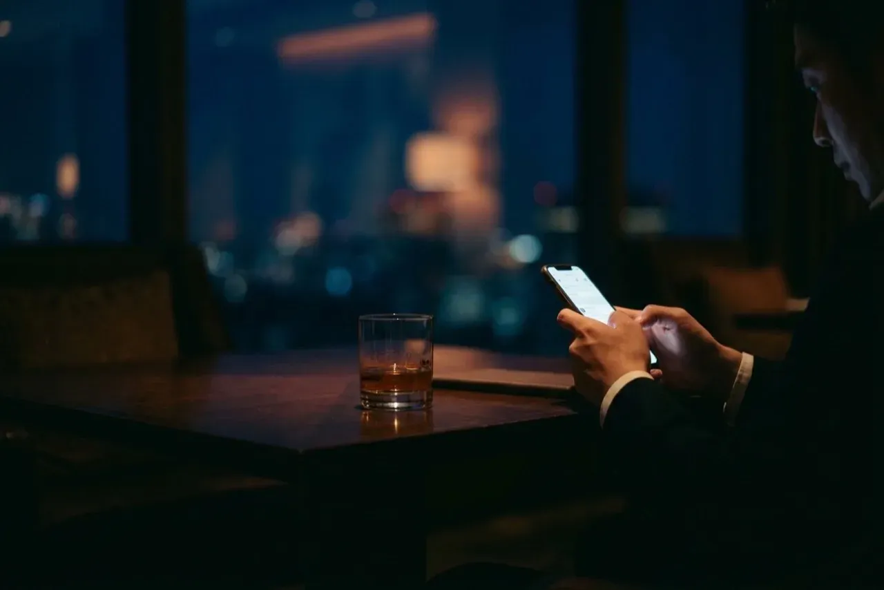 Man in a suit scrolling on his phone at night, a glass of whisky beside him against a city skyline