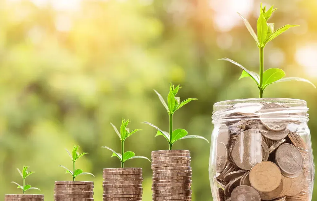 Rising stacks of coins topped with green seedlings beside a jar full of coins
