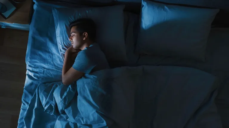 A young man asleep on his side in a dim blue bedroom, seen from above