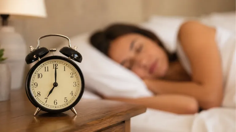 An alarm clock showing seven o'clock on a bedside table, a woman asleep behind it
