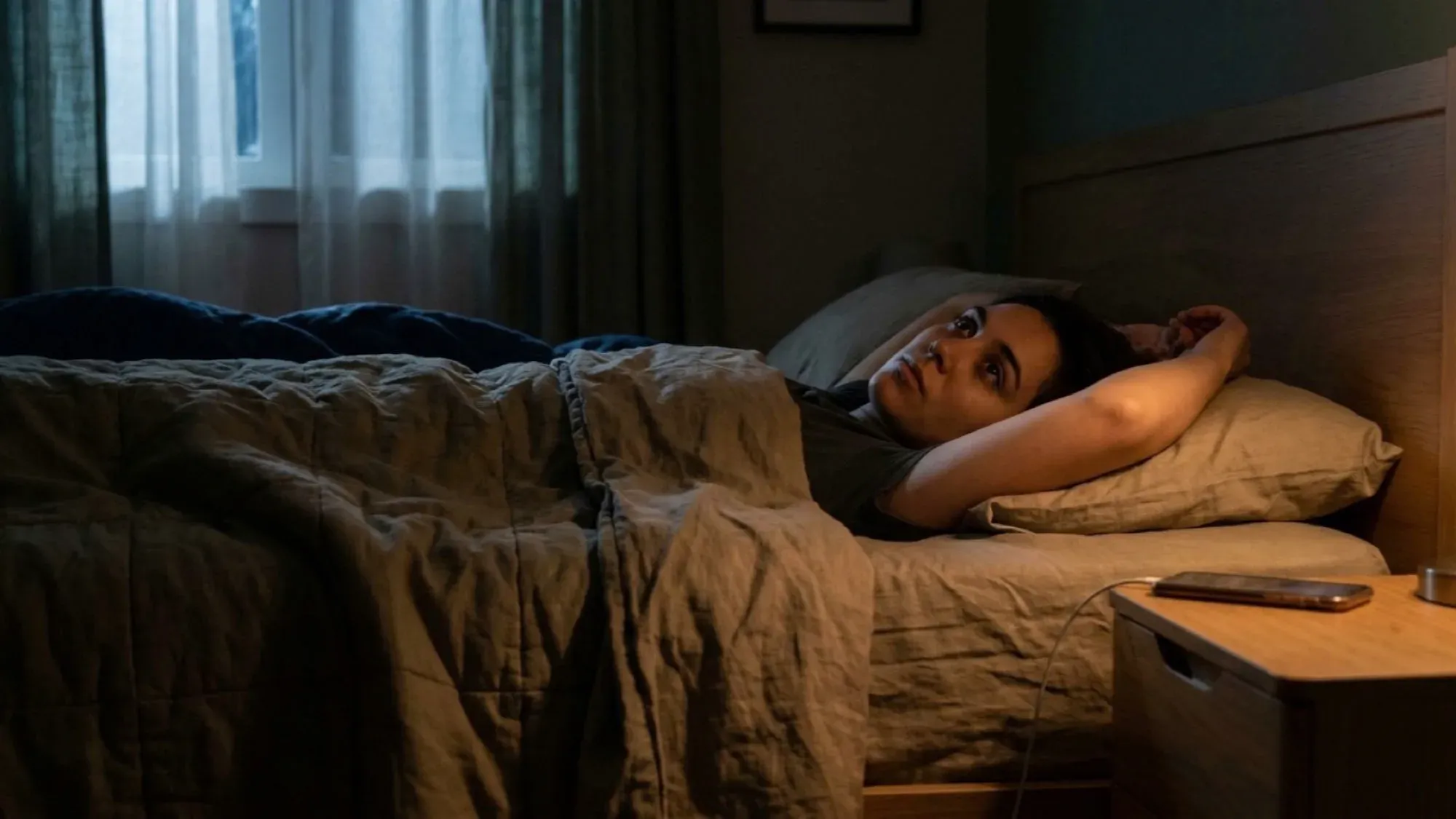 A woman lying awake in a dark bedroom, eyes open, phone charging on the bedside table