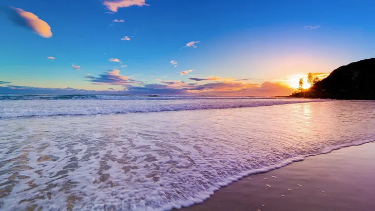 Sunrise over a beach, the sun coming up behind a headland as waves run up the sand
