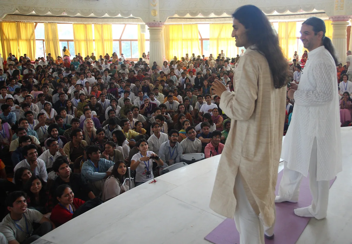 Bawa and Dinesh on stage together before a seated hall at the ashram
